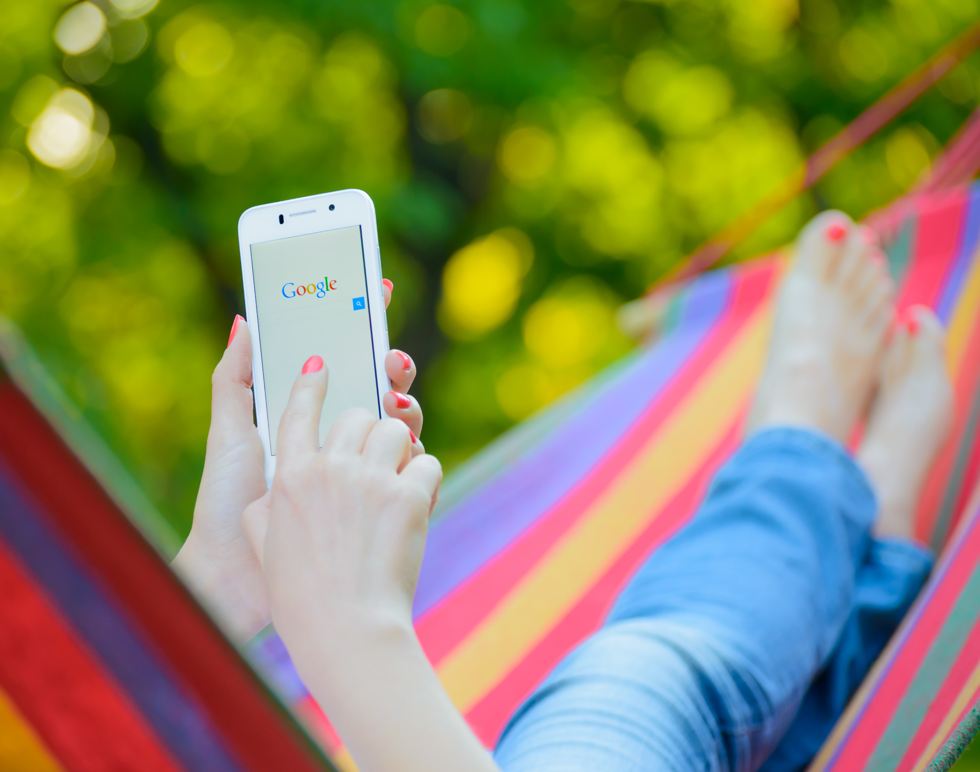 Caucasian woman with red nail polish using a cell phone searching Google for a website while laying in a rainbow-colored hammock