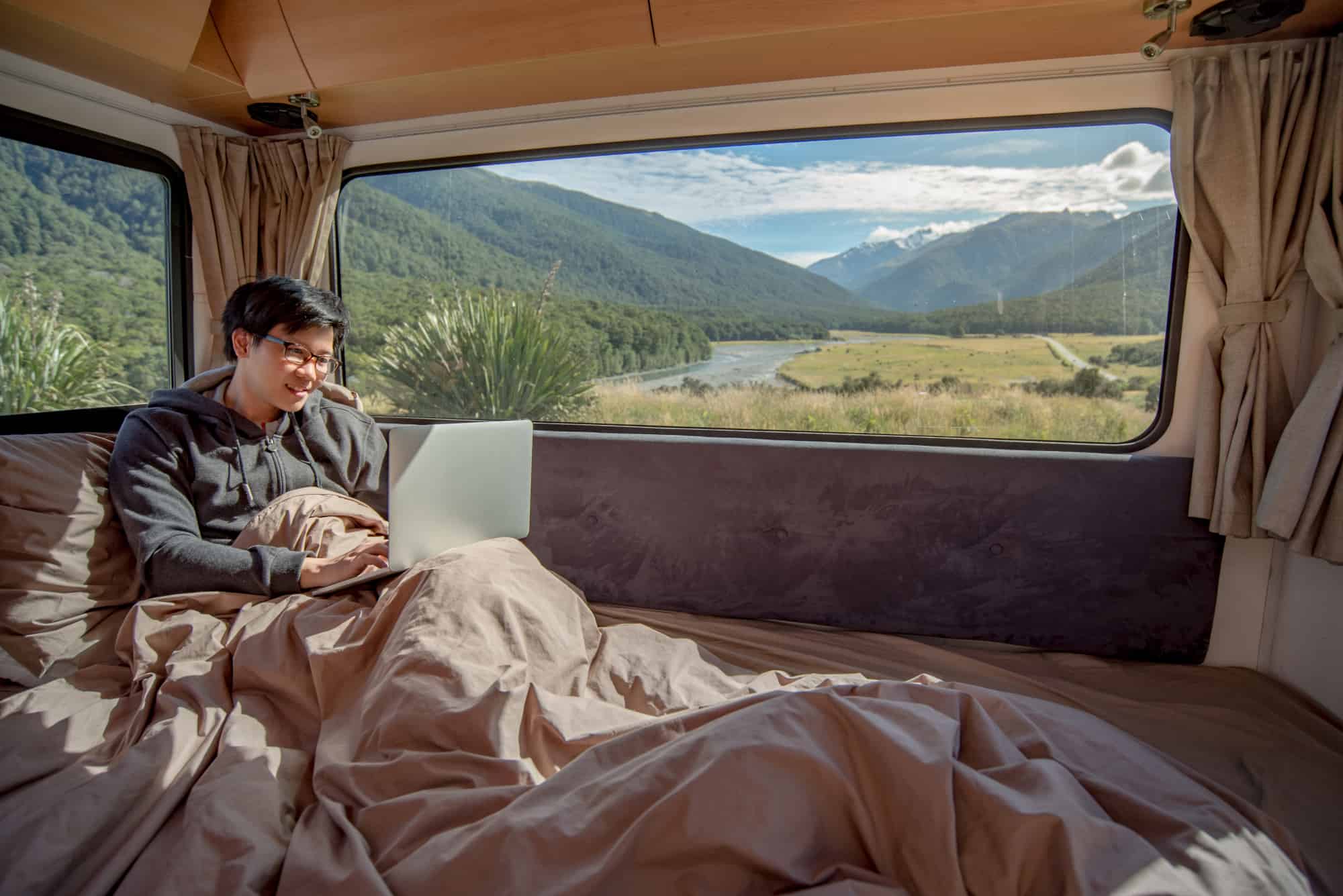 Young, male Asian digital nomad working with laptop computer on the bed with snow mountain scenic view through the window enjoying the RV lifestyle.