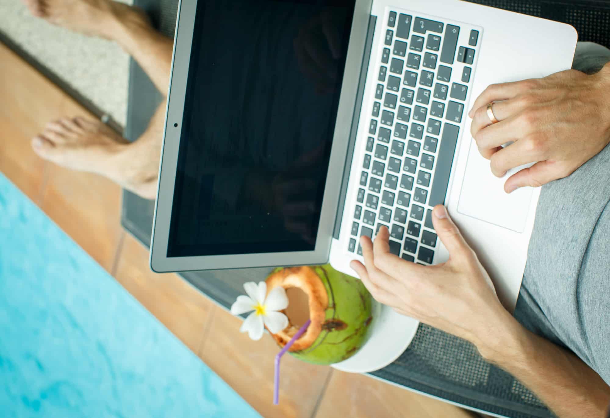 Man running payroll with Gusto on laptop sitting by the pool with a tropical drink