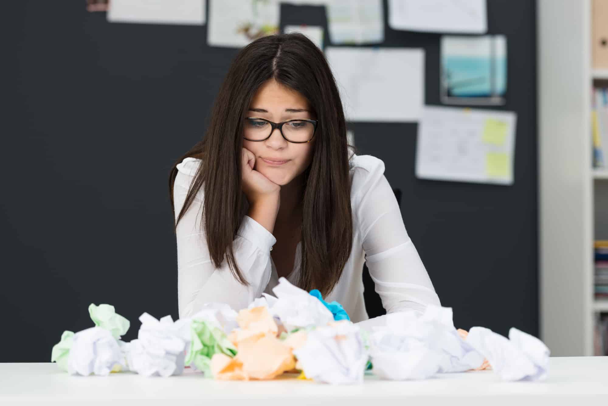 Woman wearing a white sweater and glasses working at a desk with crumbled pieces of colored paper trying to think of a name for her new small business